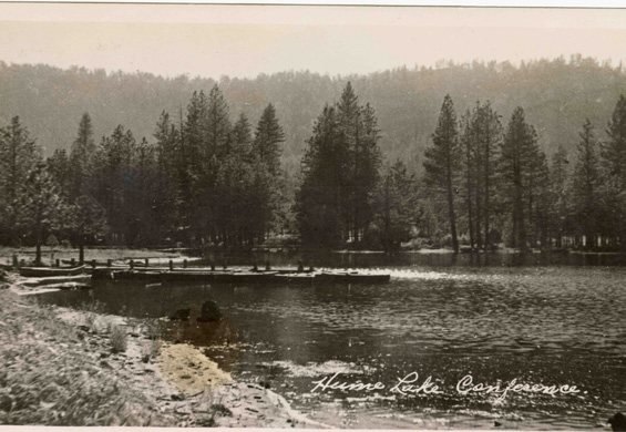 A hand-tinted 1940s postcard of Hume Lake: a dirt road curving along the shoreline, small boats at the water’s edge, pines rising to a forested ridge.
