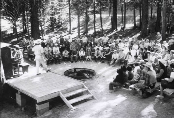 A black-and-white photograph of a large camp gathering seated on the ground around a raised wooden platform in the trees.