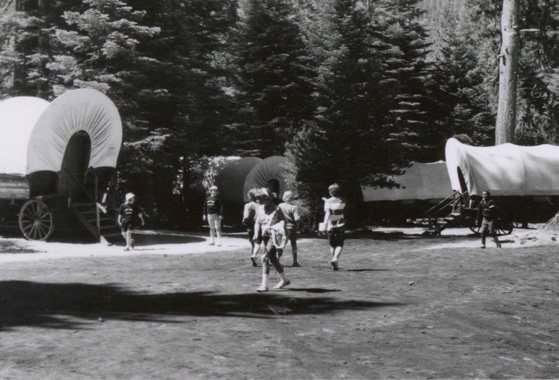 A black-and-white photograph of children walking among canvas-topped covered wagons in a clearing under tall pines.