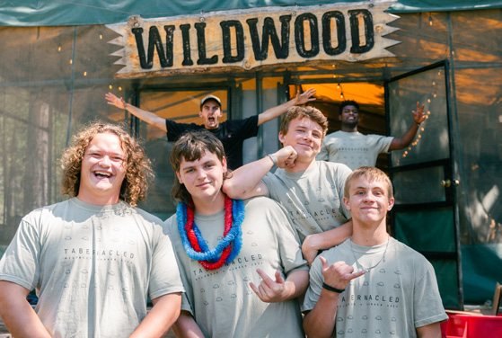 Four young men in matching grey camp shirts crowded together, laughing, beneath a hand-painted “Wildwood” banner.