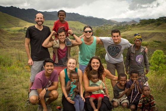 A group of adults and children posed together on a grassy hillside under a wide sky, arms around one another.