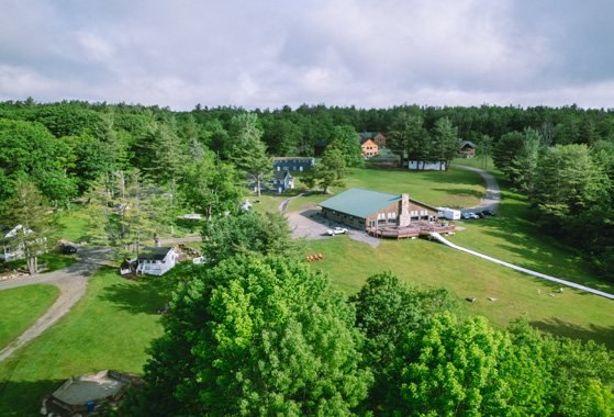 An aerial view of Hume New England: green-roofed buildings and mown fields ringed by dense hardwood forest.
