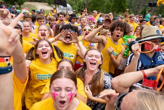 A dense crowd of campers in yellow shirts cheering with arms raised, faces painted.