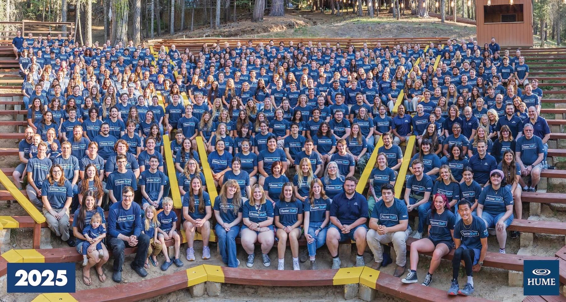 The full Hume summer staff, hundreds of people in matching Hume Staff shirts, seated together on the outdoor amphitheater benches under the pines.