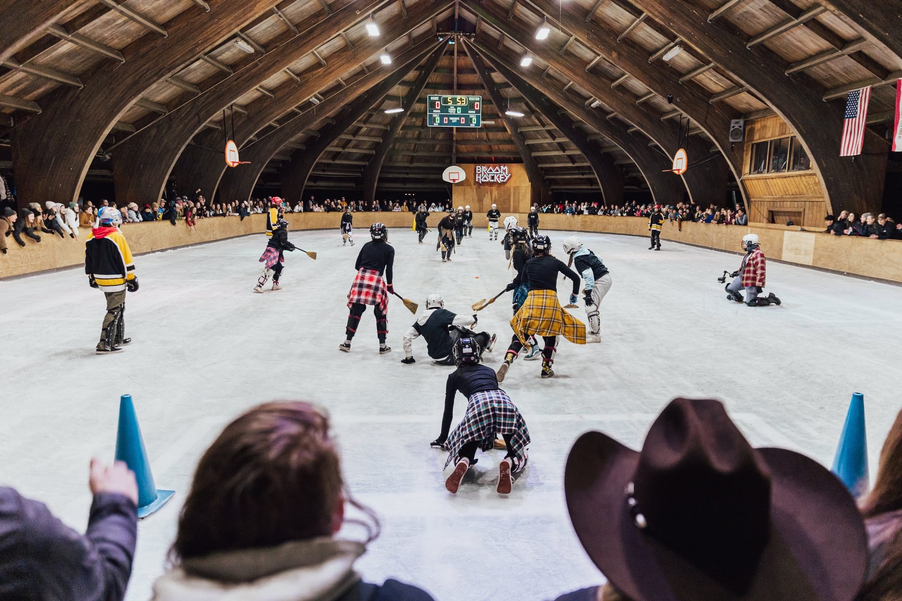 Broom hockey on the ice inside the Ark: two teams of campers in helmets and flannel chasing the ball while a packed crowd watches from the rail, under the building’s arched timber roof.