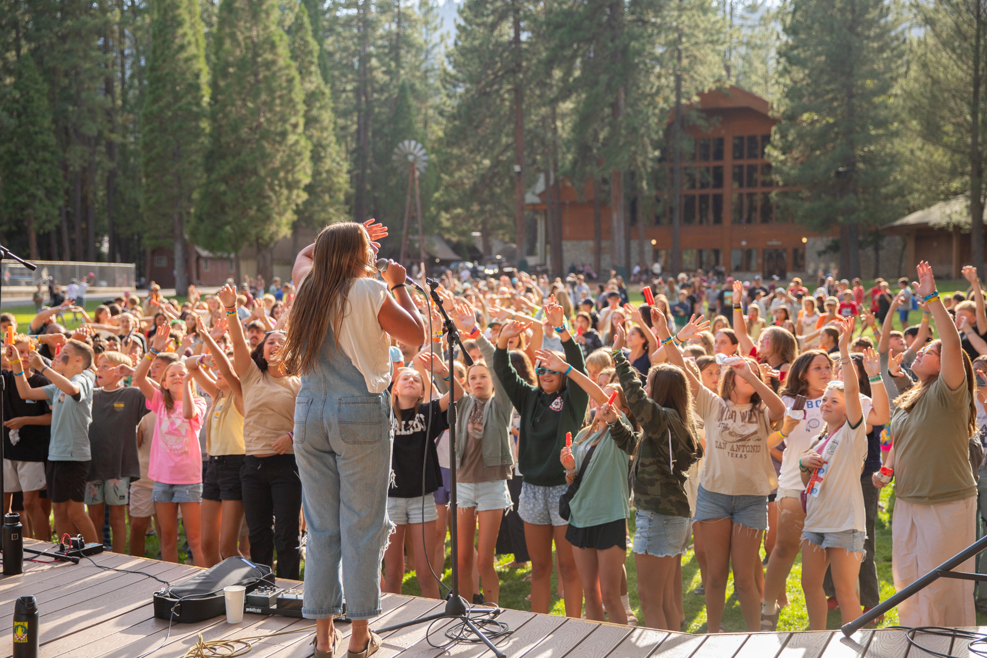 A camp worship night at Hume Lake, hundreds of campers with their hands raised as a worship leader sings from the stage.