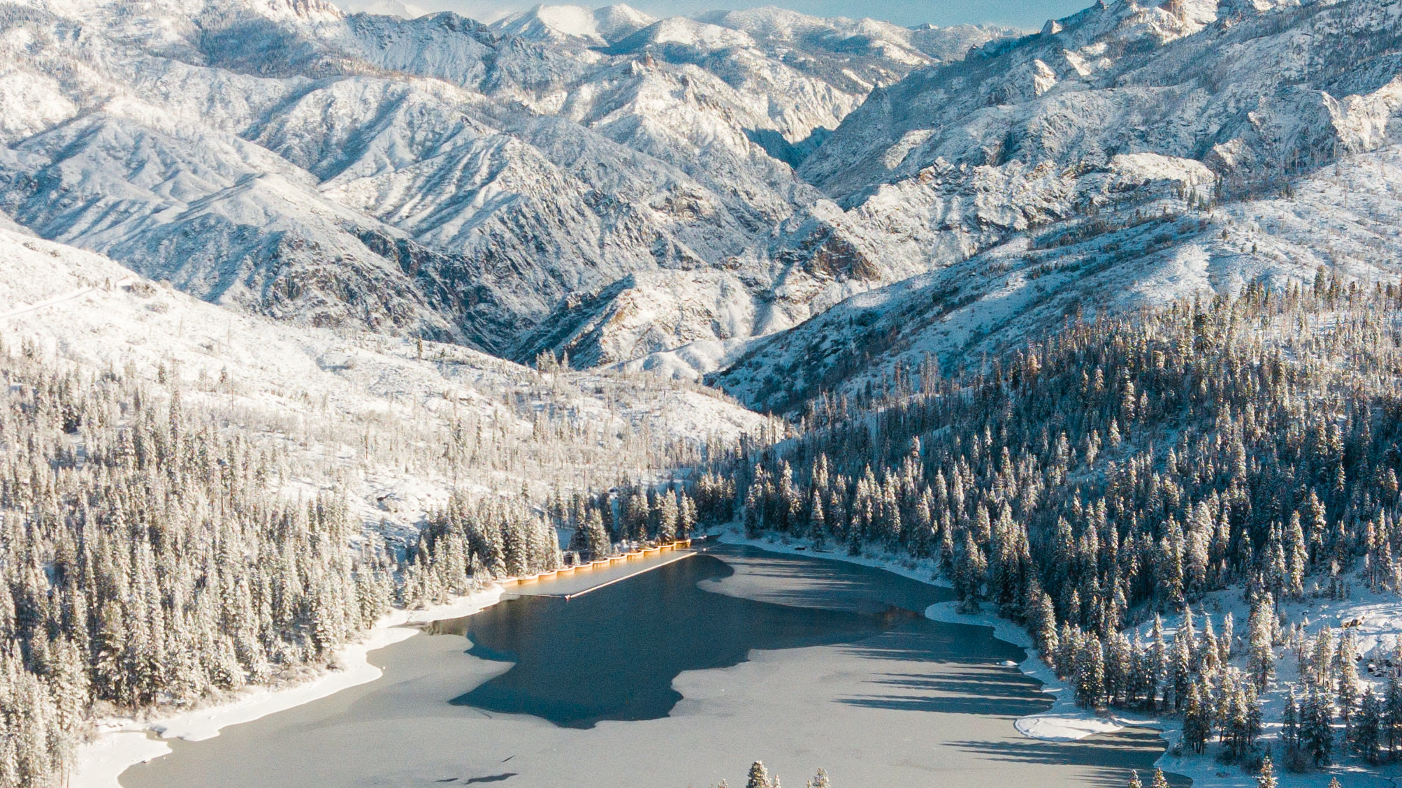 Hume Lake from the air in winter, frozen and ringed by snow-covered pines under the Sierra ridgeline.