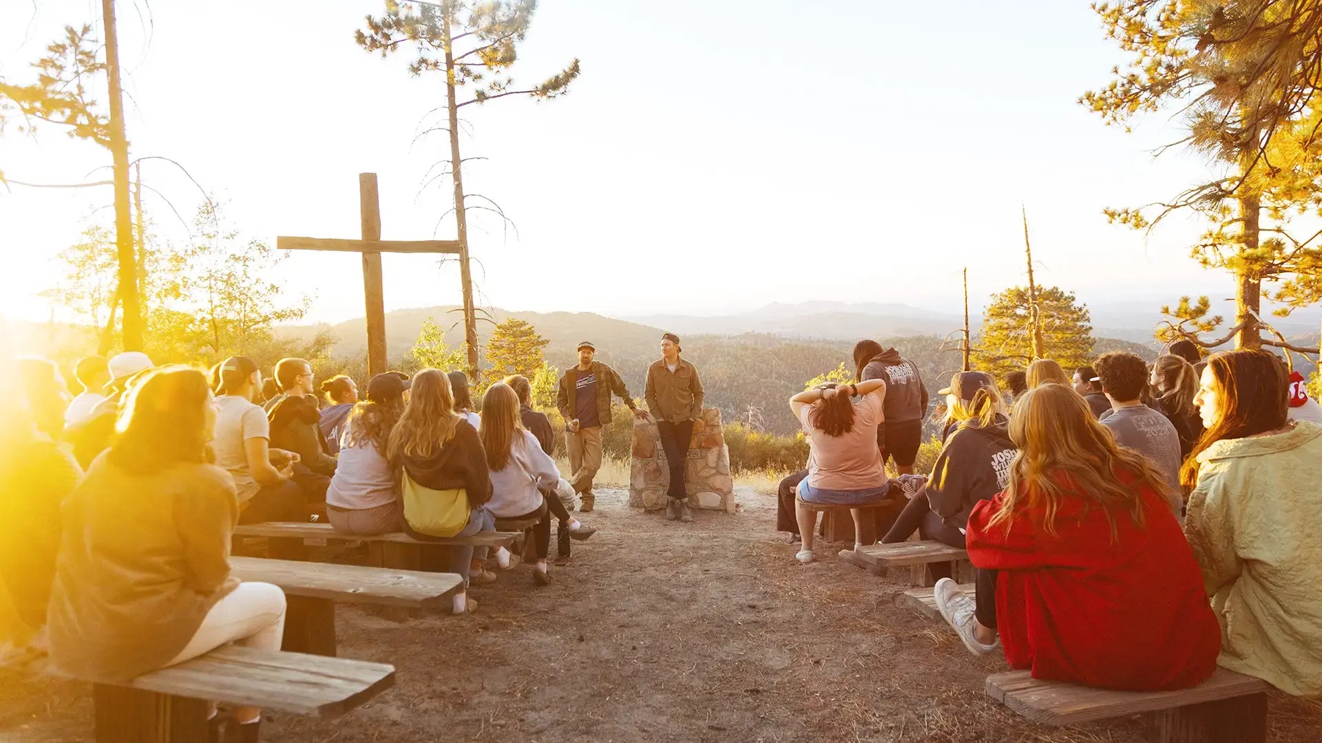 A group of young people sit on wooden benches arranged in a circle around a large wooden cross on a hilltop at sunset, with forested mountains visible in the background.