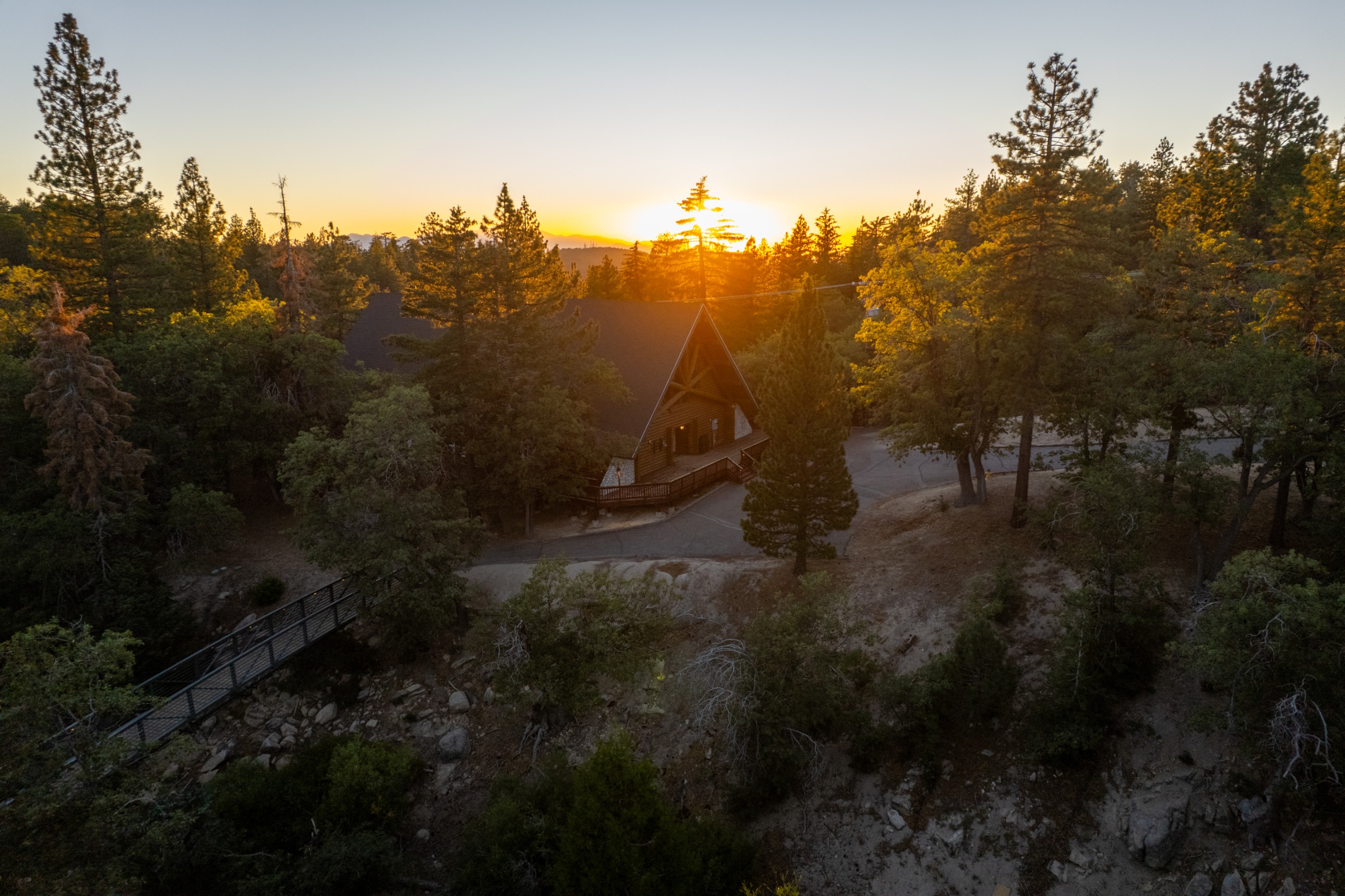 A wooden chapel with a cross on top sits nestled among tall evergreen trees at sunset, with a wooden bridge visible in the foreground.