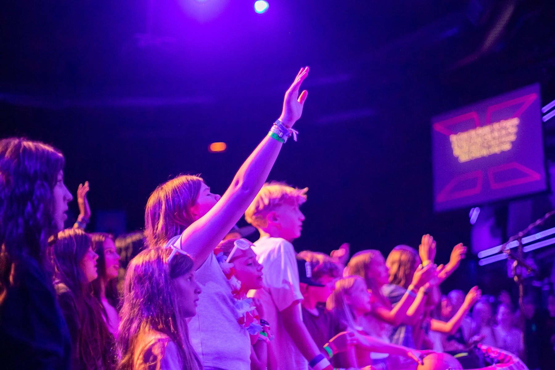 A group of young people with their hands raised dance and celebrate together under purple and pink stage lighting at an indoor event venue.