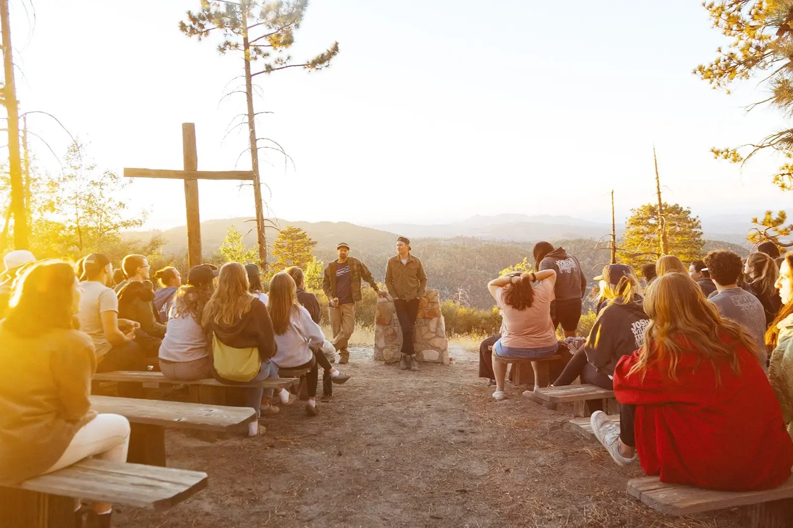 A group of young people sit on benches facing a large wooden cross on a hilltop during sunset, with forested mountains visible in the background.