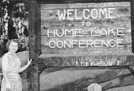 A woman in a patterned dress beside a rough-hewn wooden sign reading “Welcome to Hume Lake Conference,” in a stand of pines.