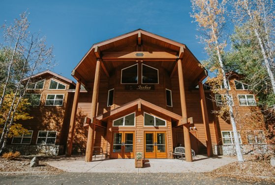A large timber-and-stone lodge with a steep shingled roof and tall windows, framed by aspens in autumn colour.
