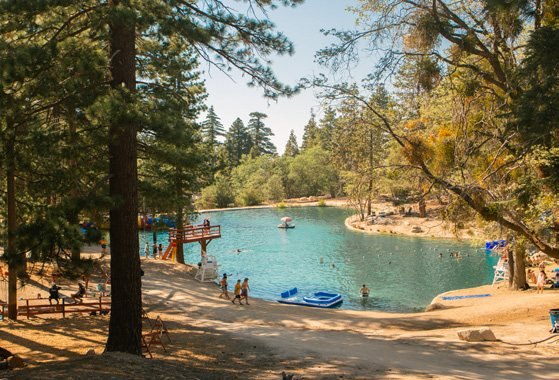 A turquoise lake edged by a sandy beach and pines, with swimmers and boats on the water.