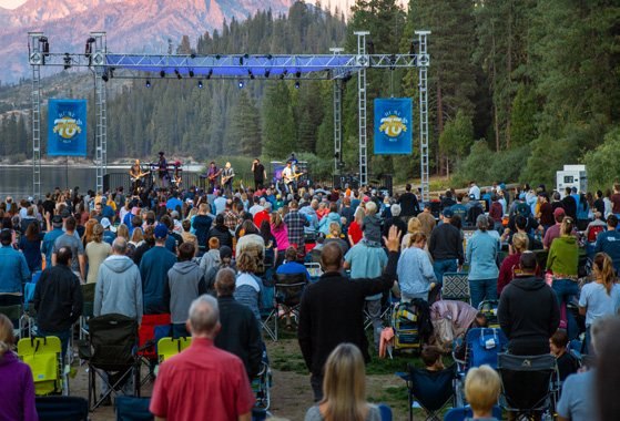 A large outdoor crowd seated and standing before a stage flanked by banners, forested mountains behind.