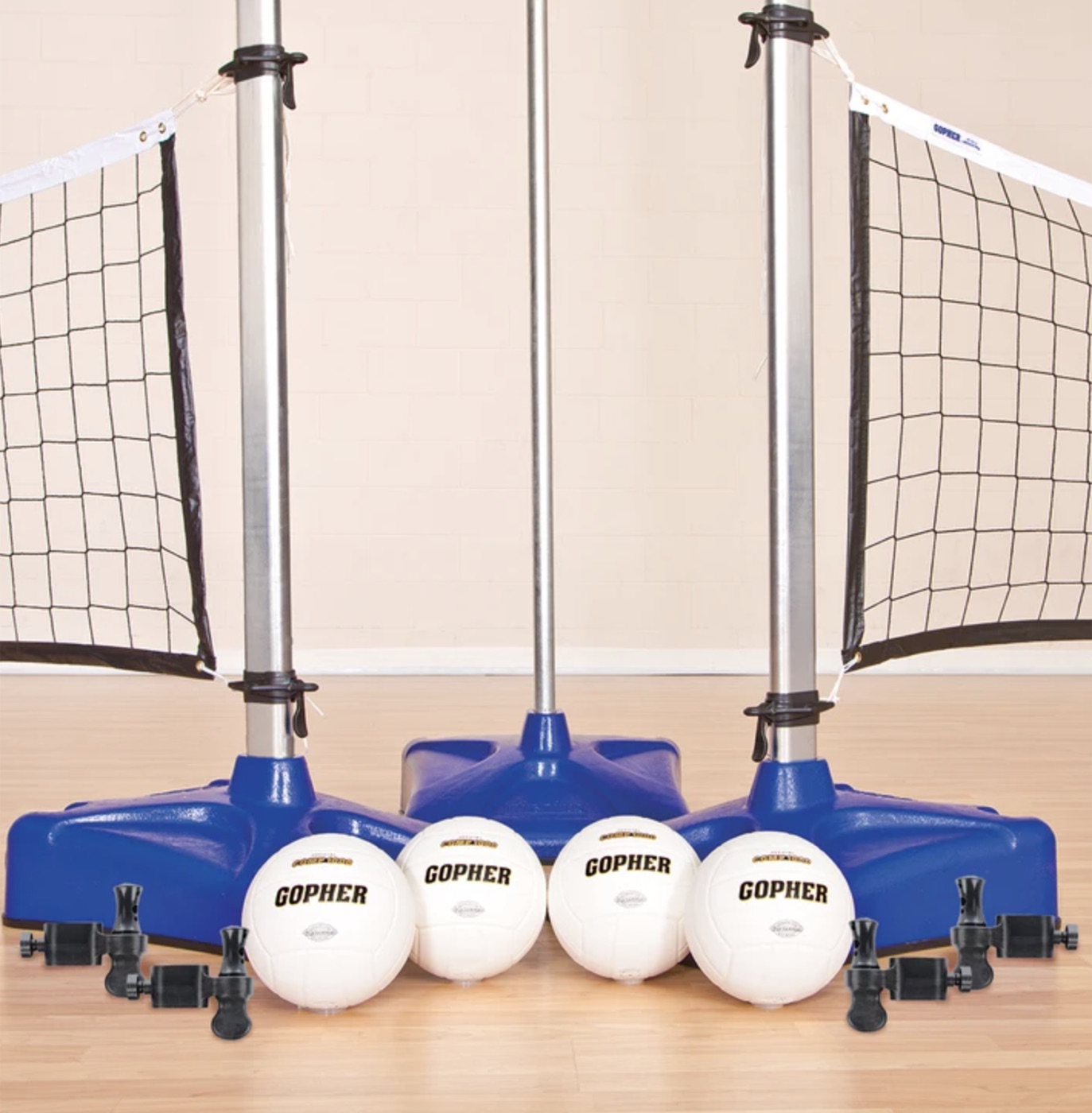 An indoor volleyball net system: three silver poles on blue bases with the net between them, and four white Gopher volleyballs on the floor in front.