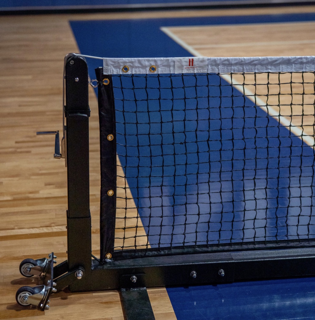 The end of a portable pickleball net on a wooden gym floor, a black post with a wheeled base holding the net, a blue court behind it.