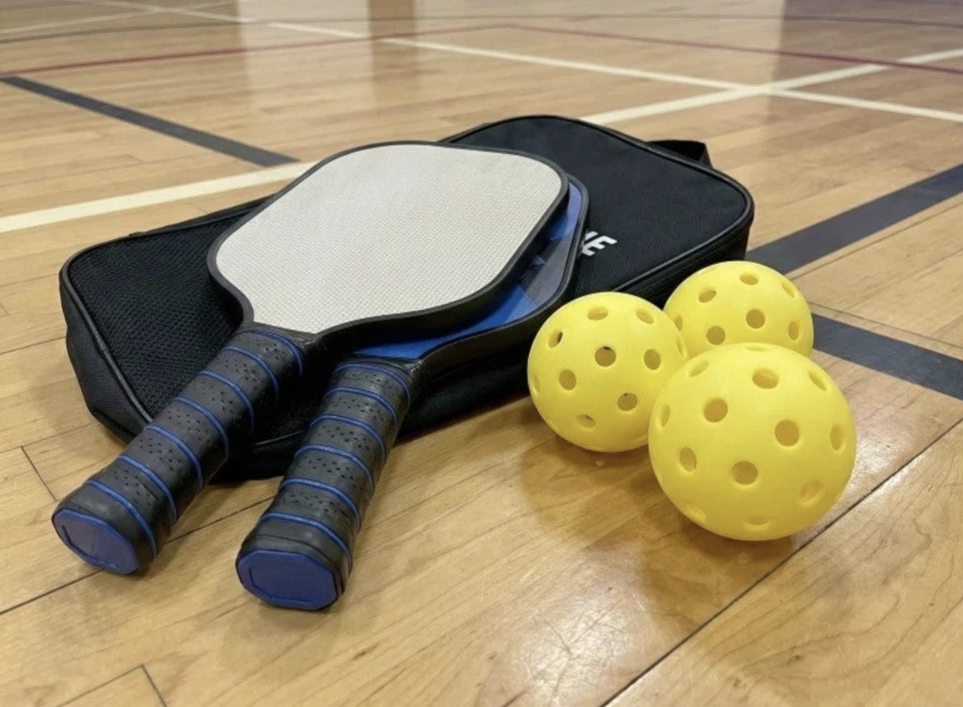 Two pickleball paddles lying on a black carrying case on a gym floor, with two yellow pickleballs beside them.